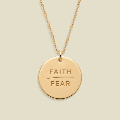 A gold disc pendant necklace engraved with the words 'FAITH' and 'FEAR' in capital letters, separated by a horizontal line, suspended from a delicate gold chain against a solid light beige background.