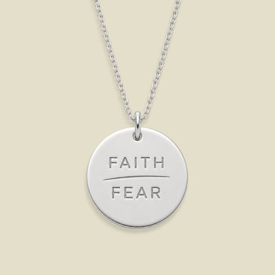 A silver round disc pendant necklace engraved with the words 'FAITH' and 'FEAR' in capital letters, separated by a horizontal line, suspended from a silver chain against a solid light beige background.