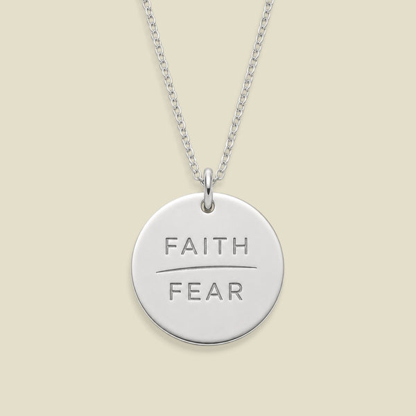 A silver round disc pendant necklace engraved with the words 'FAITH' and 'FEAR' in capital letters, separated by a horizontal line, suspended from a silver chain against a solid light beige background.