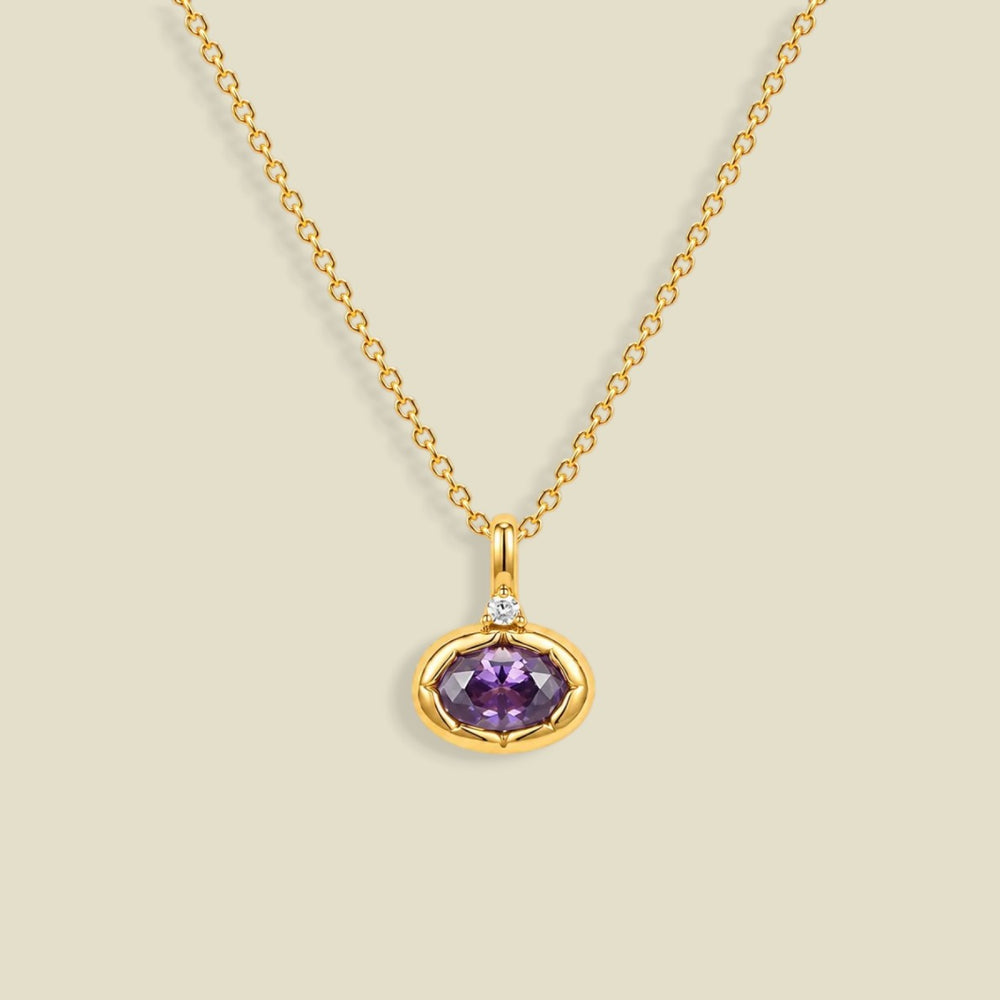 A gold chain necklace with a pendant featuring a bezel-set oval purple CZ gemstone and a small clear accent stone above it, shown on a light beige background.