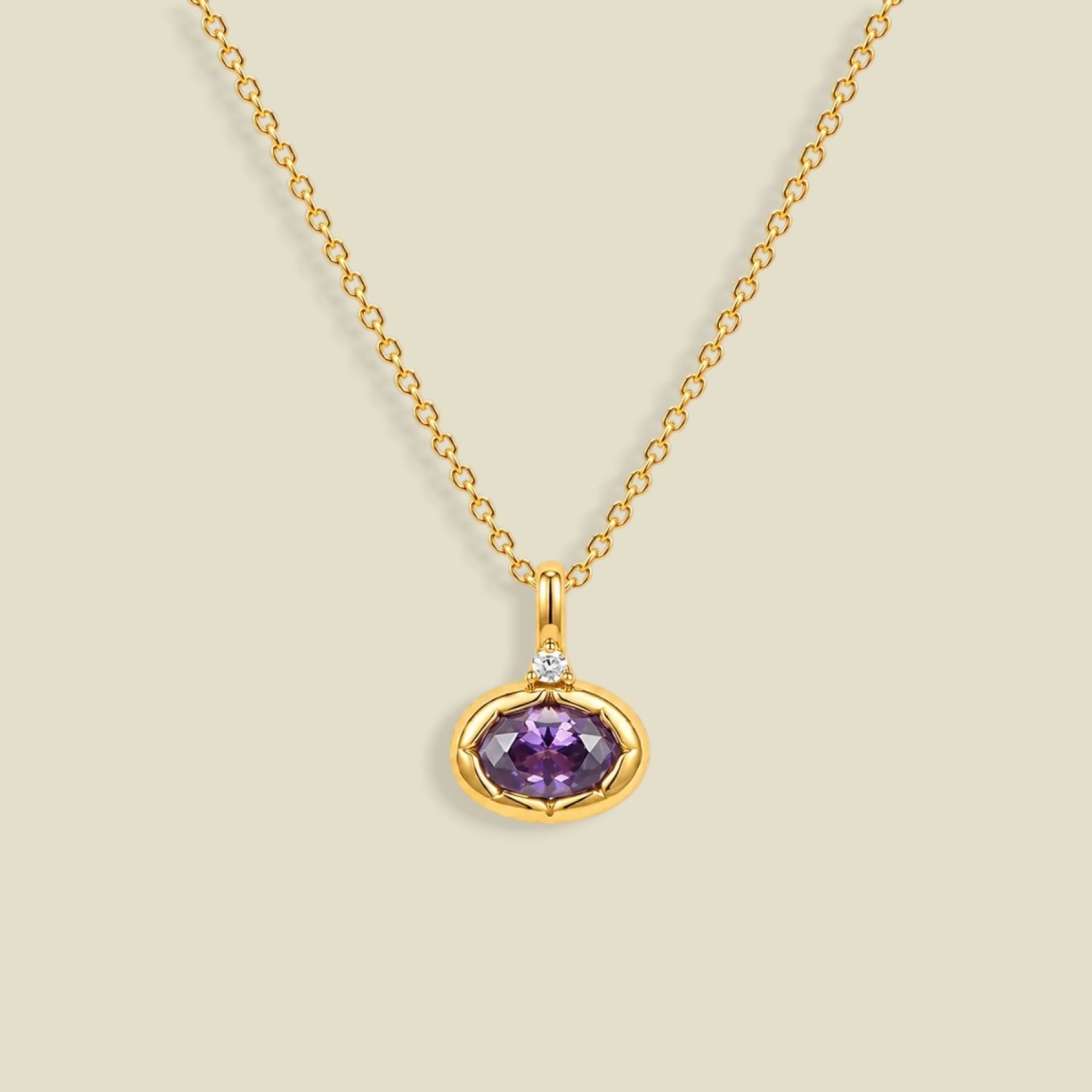 A gold chain necklace with a pendant featuring a bezel-set oval purple CZ gemstone and a small clear accent stone above it, shown on a light beige background.