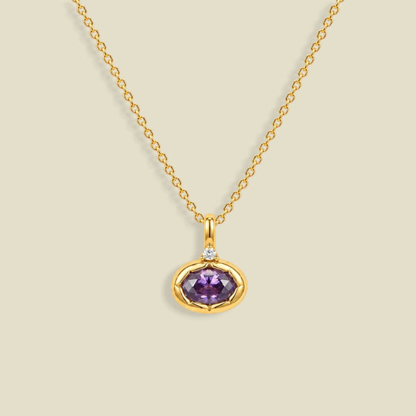 A gold chain necklace with a pendant featuring a bezel-set oval purple CZ gemstone and a small clear accent stone above it, shown on a light beige background.