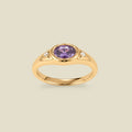 A gold signet ring with a purple center oval bezel-set gemstone, and two small clear accent stones on either side, set against a solid beige background.