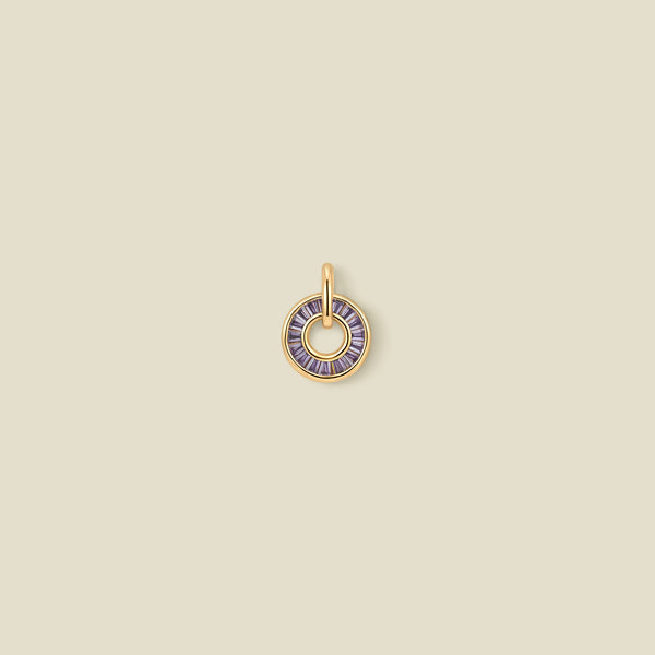 A circular gold pendant featuring purple baguette-cut stones around a hollow center, displayed against a solid light beige background.