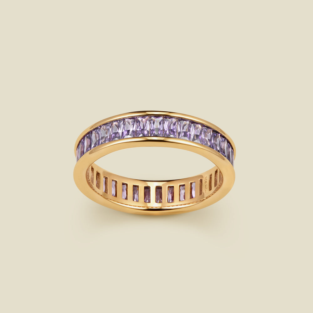 A gold eternity ring featuring a channel-set row of baguette-cut light purple gemstones on the outer band, shown against a solid light beige background.