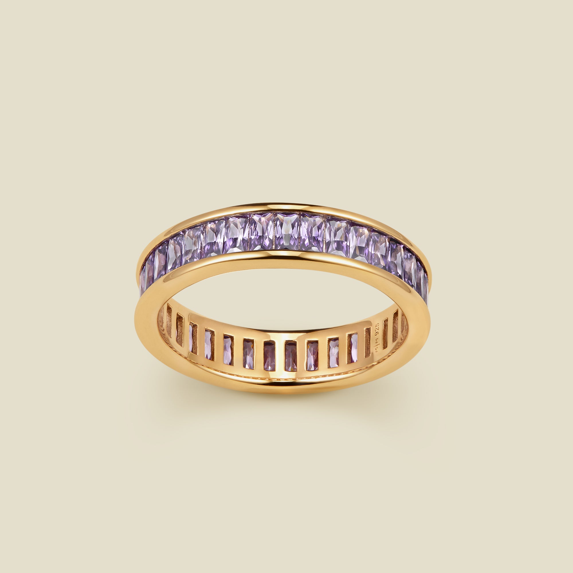 A gold eternity ring featuring a channel-set row of baguette-cut light purple gemstones on the outer band, shown against a solid light beige background.