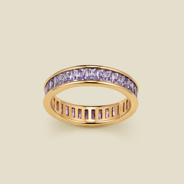 A gold eternity ring featuring a channel-set row of baguette-cut light purple gemstones on the outer band, shown against a solid light beige background.