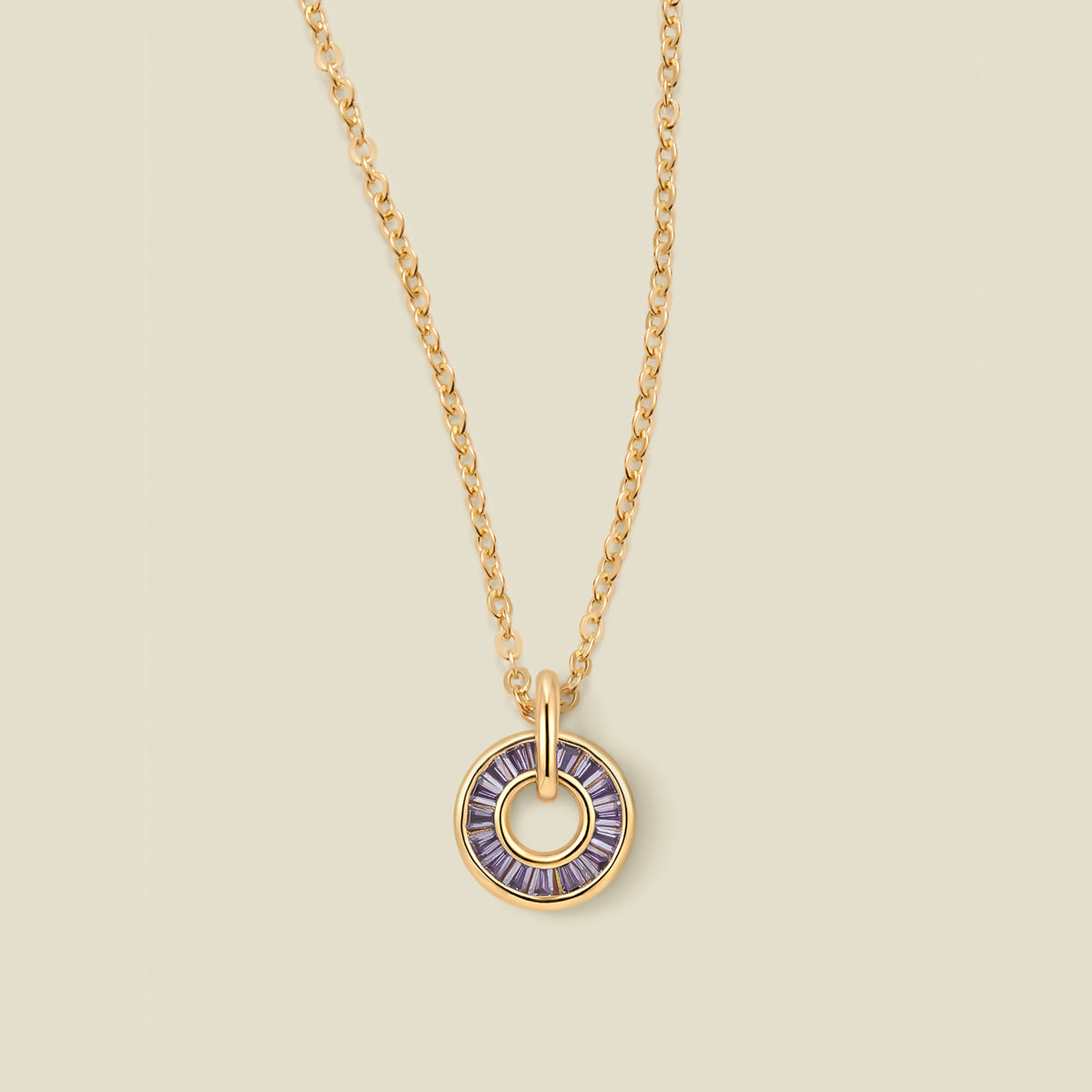 A gold chain necklace with a circular pendant featuring purple baguette-cut stones around a hollow center, displayed against a solid light beige background.