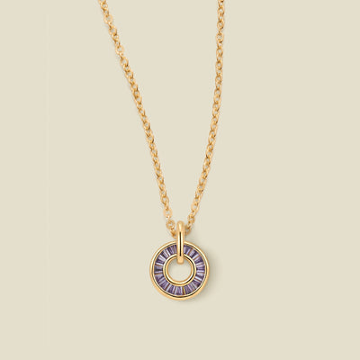 A gold chain necklace with a circular pendant featuring purple baguette-cut stones around a hollow center, displayed against a solid light beige background.