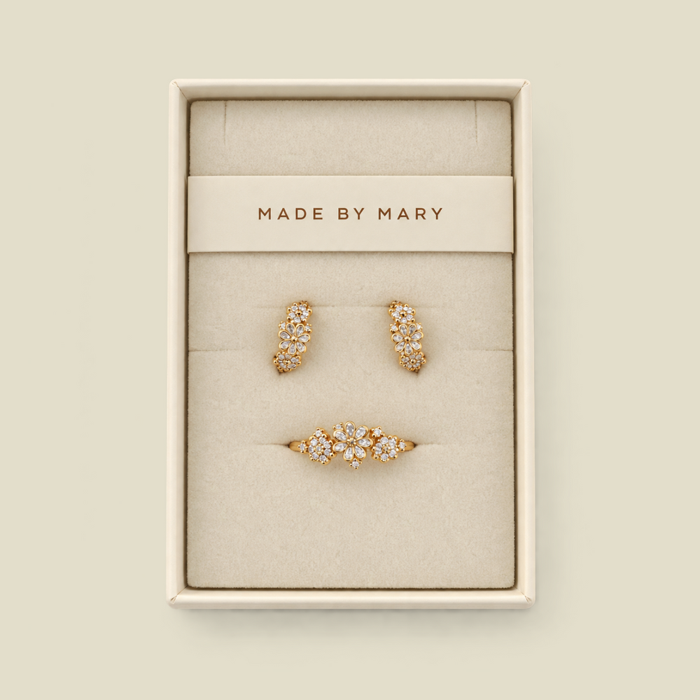 A three-piece gold jewelry set by MADE BY MARY, including two huggie hoop earrings and a flower cluster ring, displayed in a cream-colored jewelry box on a matching beige background.