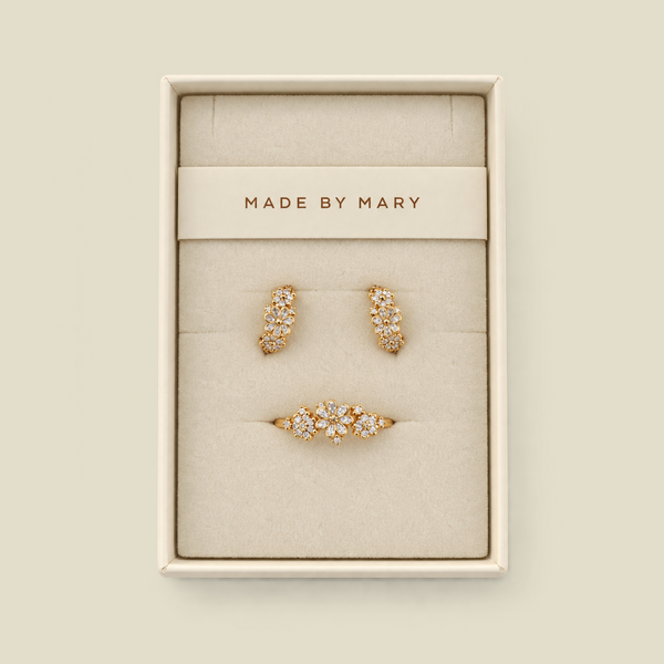 A three-piece gold jewelry set by MADE BY MARY, including two huggie hoop earrings and a flower cluster ring, displayed in a cream-colored jewelry box on a matching beige background.