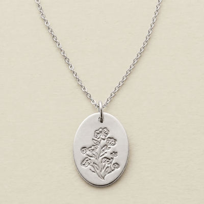 A silver oval pendant necklace featuring an engraved floral design, suspended from a delicate silver chain against a light beige textured background.