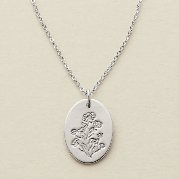A silver oval pendant necklace featuring an engraved floral design, suspended from a delicate silver chain against a light beige textured background.