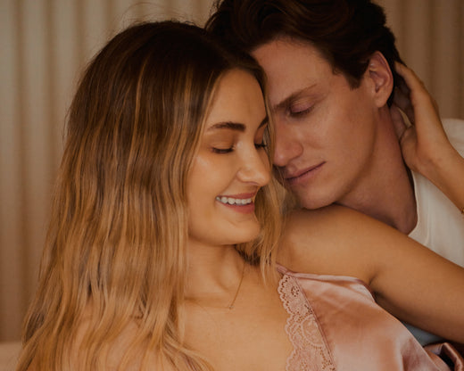 A close-up lifestyle shot of a smiling woman with long blonde hair wearing a pink satin camisole with lace trim, embraced by a man with dark hair in a warm, softly lit indoor setting.