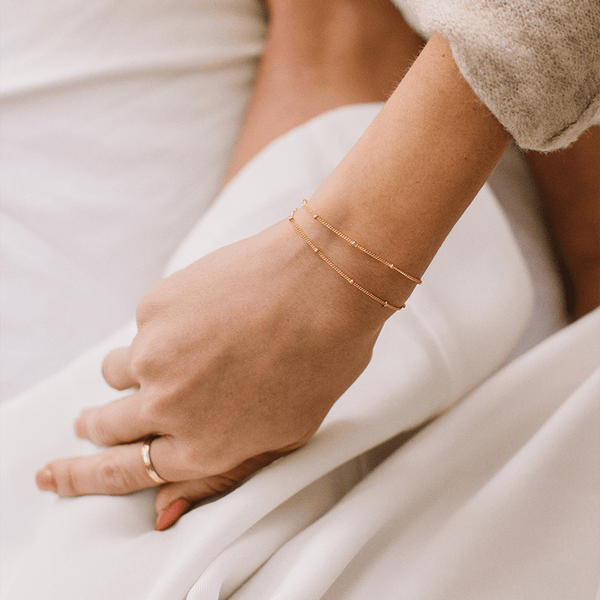 A person's wrist wearing two thin gold chain bracelets, each with stationary round polished beads.