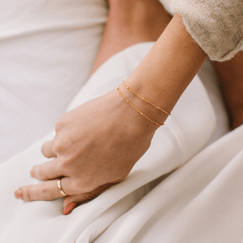 A person's wrist wearing two thin gold chain bracelets, each with stationary round polished beads.