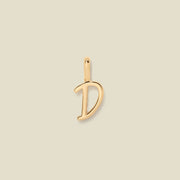 A polished gold initial pendant charm shaped like the letter 'D' in a cursive script, centered on a solid light beige background.