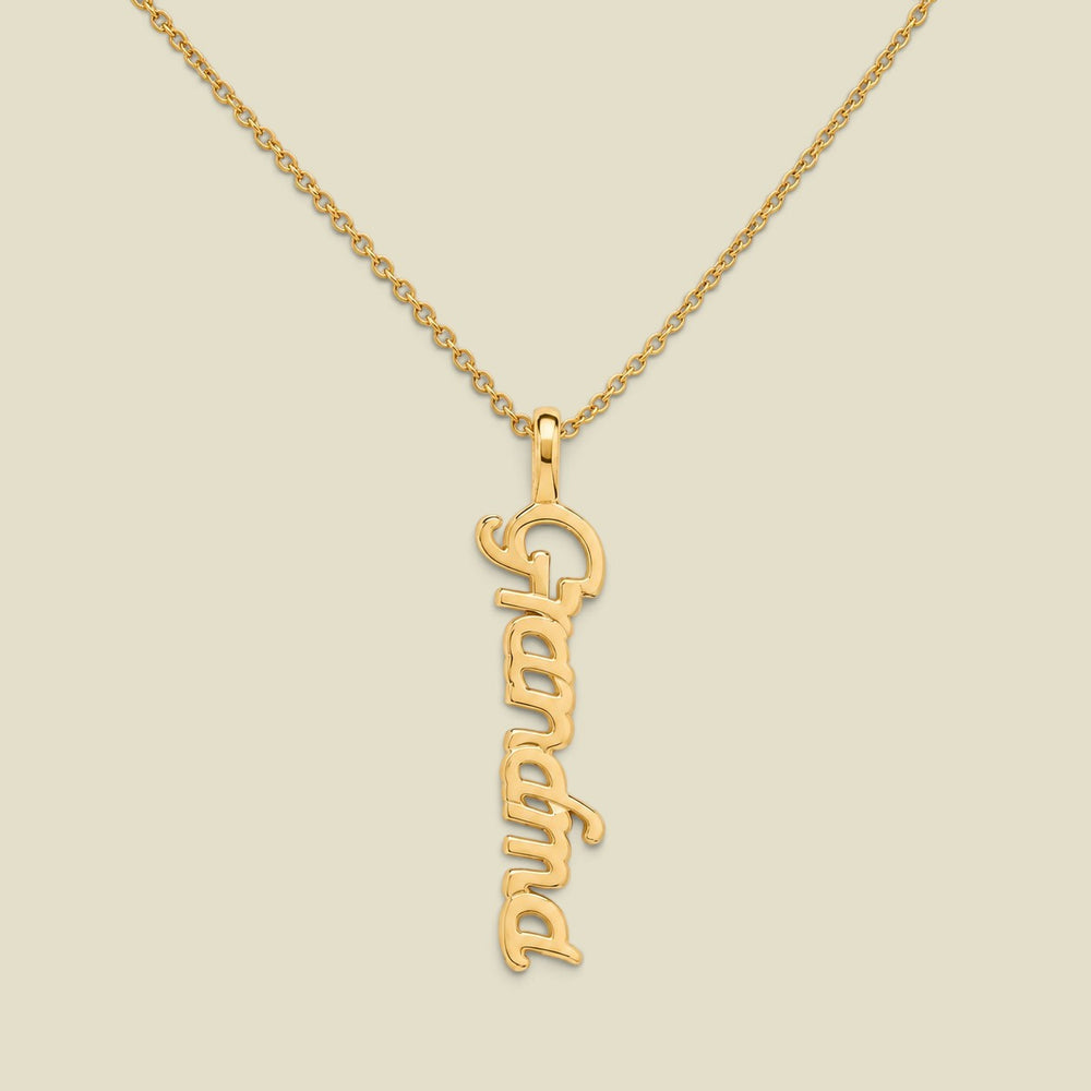 A gold script nameplate necklace spelling 'Grandma'  hanging vertically on a delicate gold chain, presented on a solid light beige background.