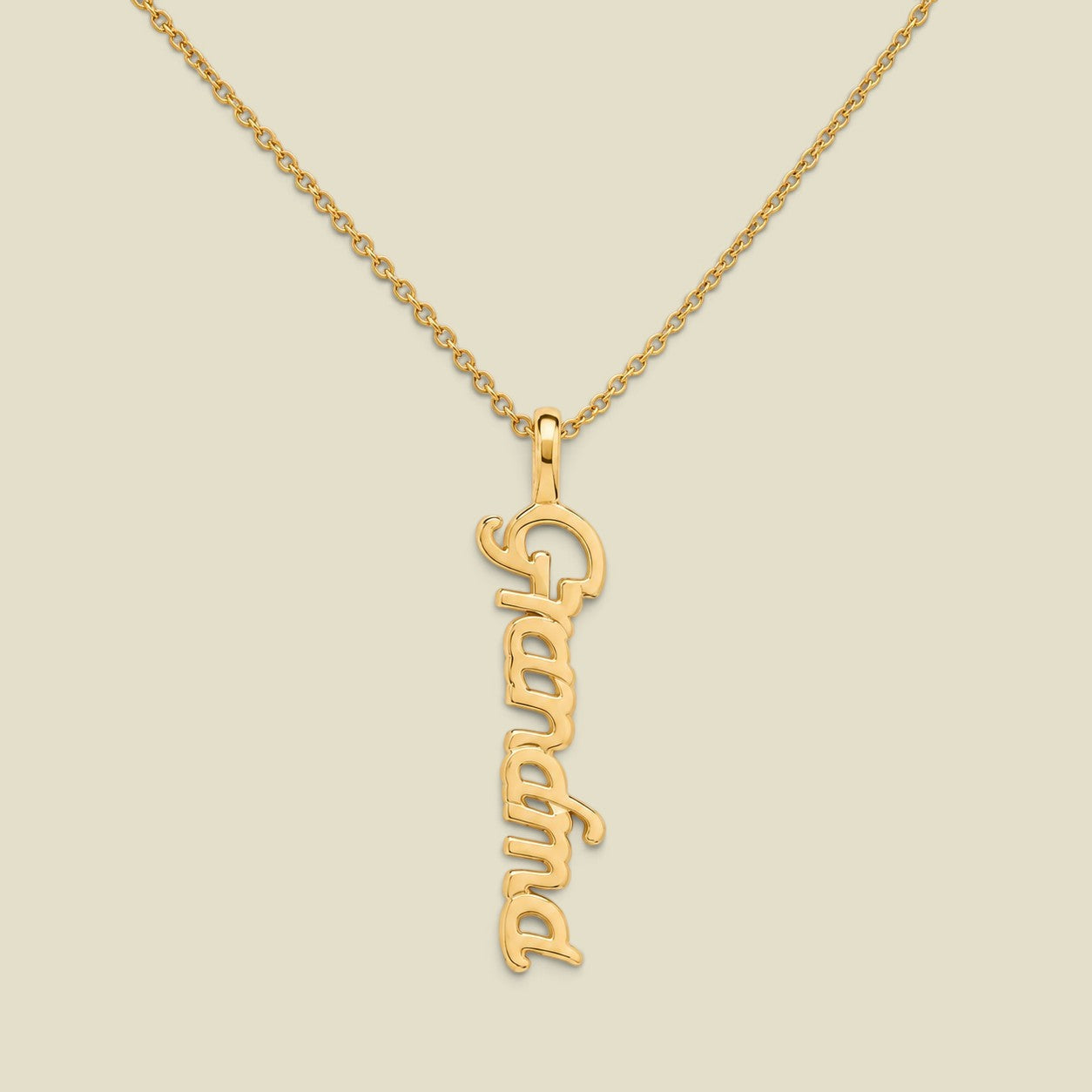A gold script nameplate necklace spelling 'Grandma'  hanging vertically on a delicate gold chain, presented on a solid light beige background.