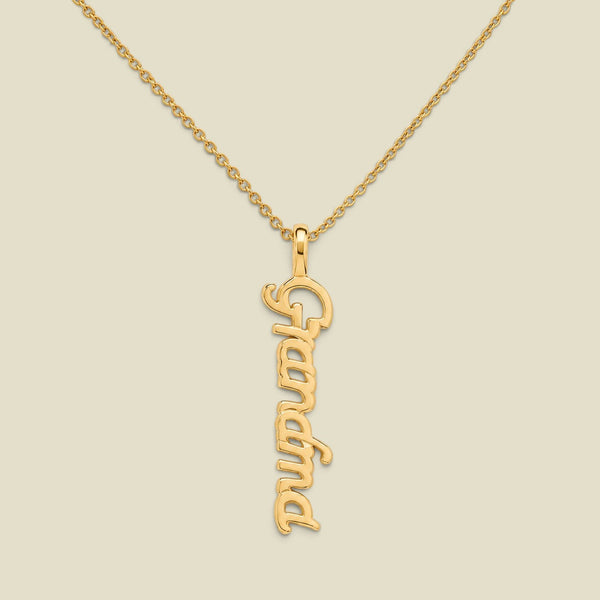 A gold script nameplate necklace spelling 'Grandma'  hanging vertically on a delicate gold chain, presented on a solid light beige background.