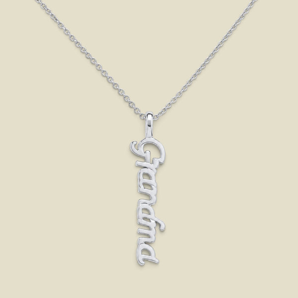 A silver script nameplate necklace spelling 'Grandma'  hanging vertically on a delicate silver chain, presented on a solid light beige background.