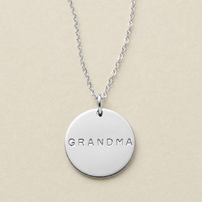 A silver round disc pendant necklace engraved with the word 'GRANDMA' in uppercase letters, hanging from a delicate silver chain against a solid beige background.