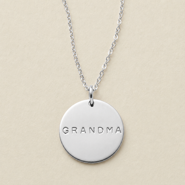 A silver round disc pendant necklace engraved with the word 'GRANDMA' in uppercase letters, hanging from a delicate silver chain against a solid beige background.