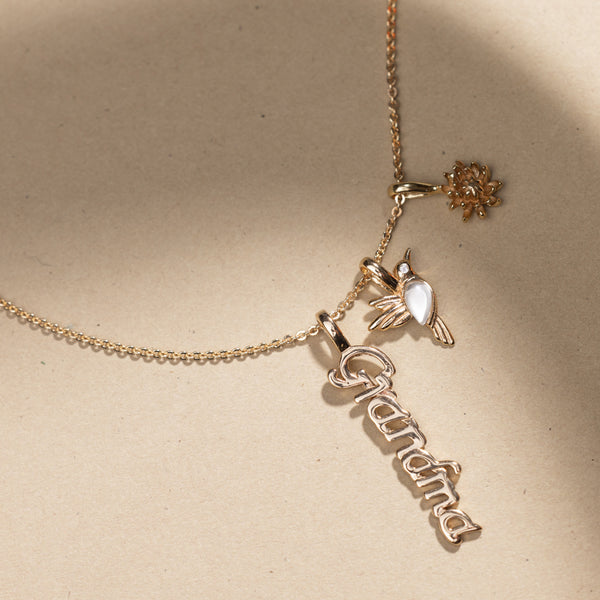 A delicate silver chain necklace featuring a small bird charm with a white gemstone, a nameplate charm spelling 'Grandma', and a small flower charm, displayed on a textured beige background.