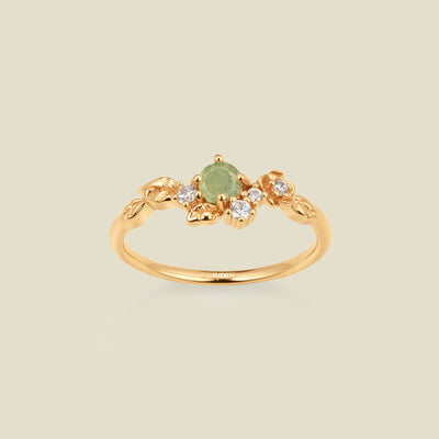 Gold ring with floral embellishments and green and clear prong-set stones against a neutral background.
