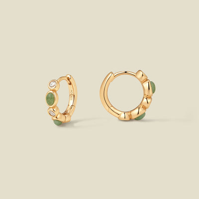 Pair of gold hoop earrings with green and clear bezel set stones against a neutral background.