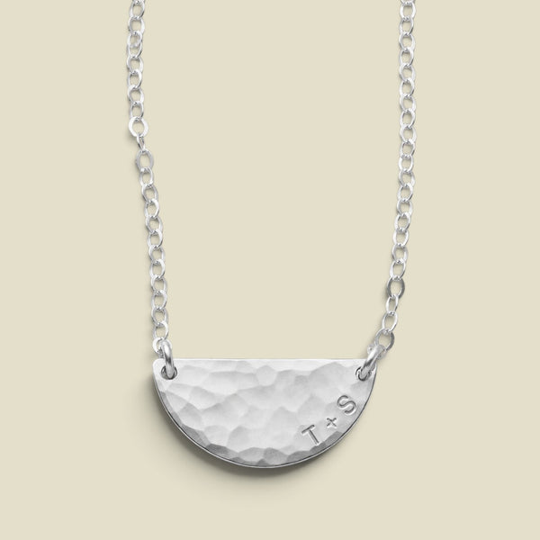 A silver necklace featuring a hammered semi-circular pendant with engraved initials 'T+S', suspended from both ends to a delicate link chain against a solid beige background.