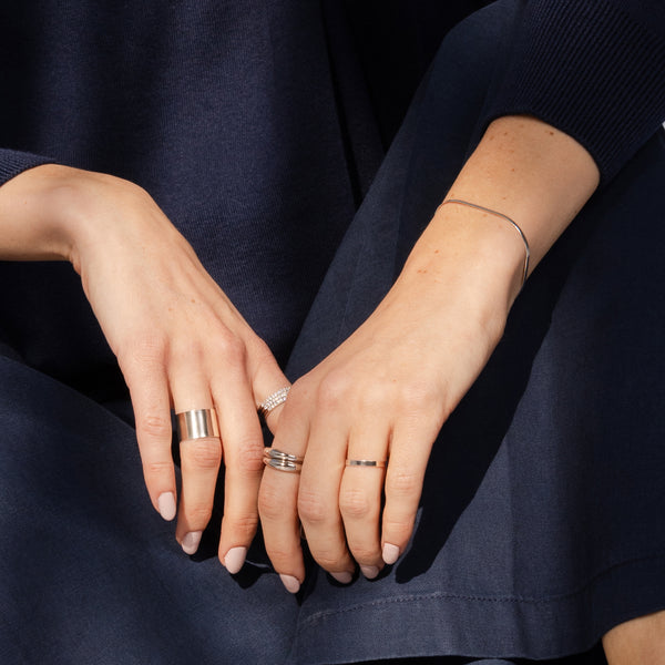 Close-up of a person's hands resting on their lap, wearing several minimalist silver rings and a delicate silver bracelet against a dark navy blue knit top and skirt.