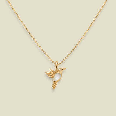 A gold chain necklace with a small bird pendant featuring a white gemstone bodice, displayed against a solid light beige background.