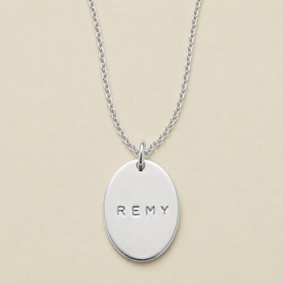 A silver oval pendant necklace engraved with the name 'REMY', displayed on a delicate silver chain against a solid beige background.