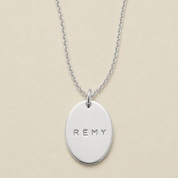 A silver oval pendant necklace engraved with the name 'REMY', displayed on a delicate silver chain against a solid beige background.