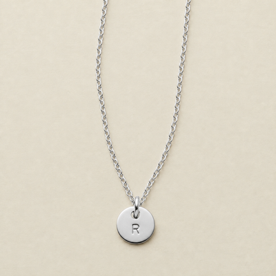 A silver chain necklace with a round disc pendant engraved with the letter 'R', displayed on a light beige background.
