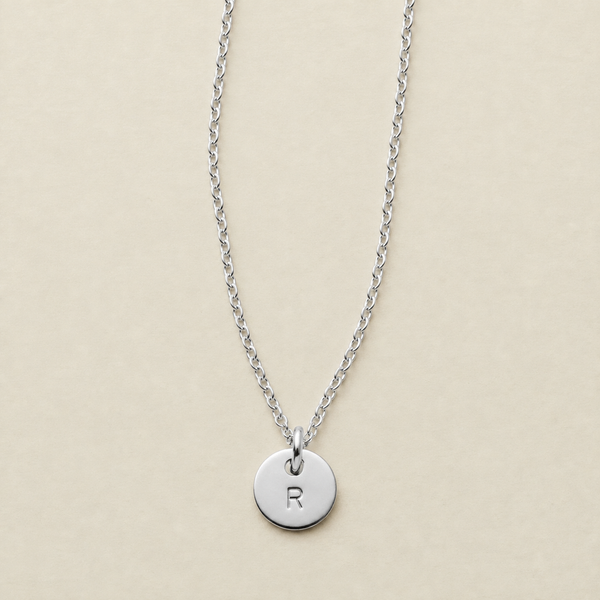 A silver chain necklace with a round disc pendant engraved with the letter 'R', displayed on a light beige background.