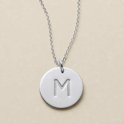 A silver round disc pendant necklace engraved with the letter 'M', hanging from a delicate silver chain against a solid beige background.