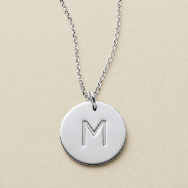 A silver round disc pendant necklace engraved with the letter 'M', hanging from a delicate silver chain against a solid beige background.