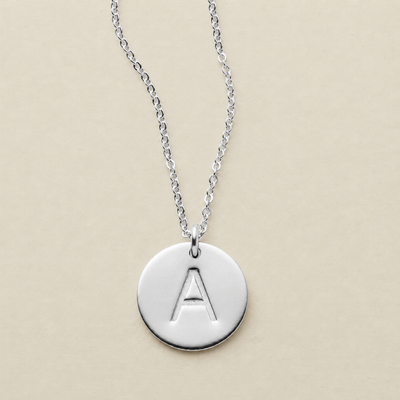 A silver round disc pendant necklace with an engraved capital letter 'A', hanging from a delicate silver chain against a solid beige background.
