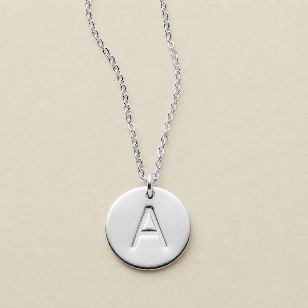 A silver round disc pendant necklace with an engraved capital letter 'A', hanging from a delicate silver chain against a solid beige background.