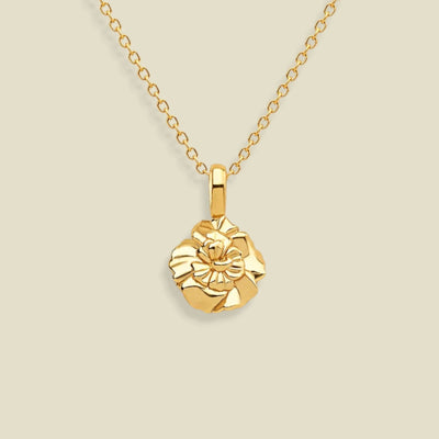 A gold necklace with a small, textured carnation flower pendant, displayed against a solid light beige background.