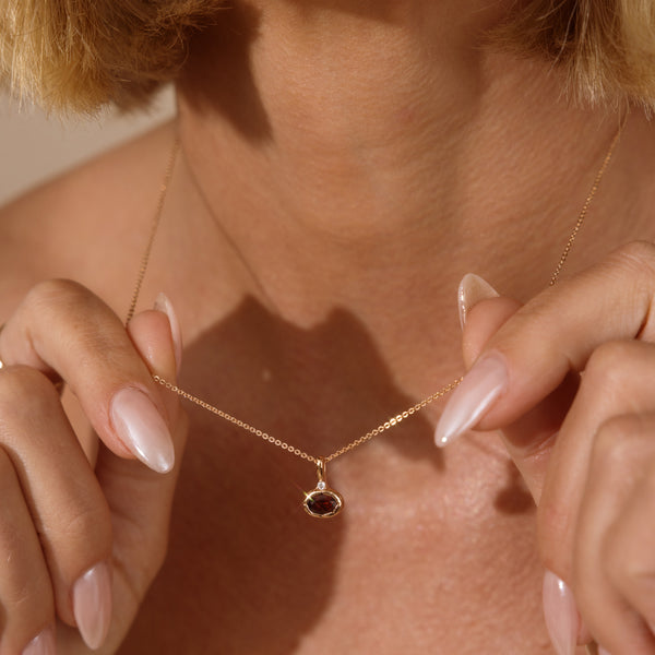A dainty gold pendant necklace with an oval bezel set red gemstone charm, with a small clear cz above it on a person's neck.