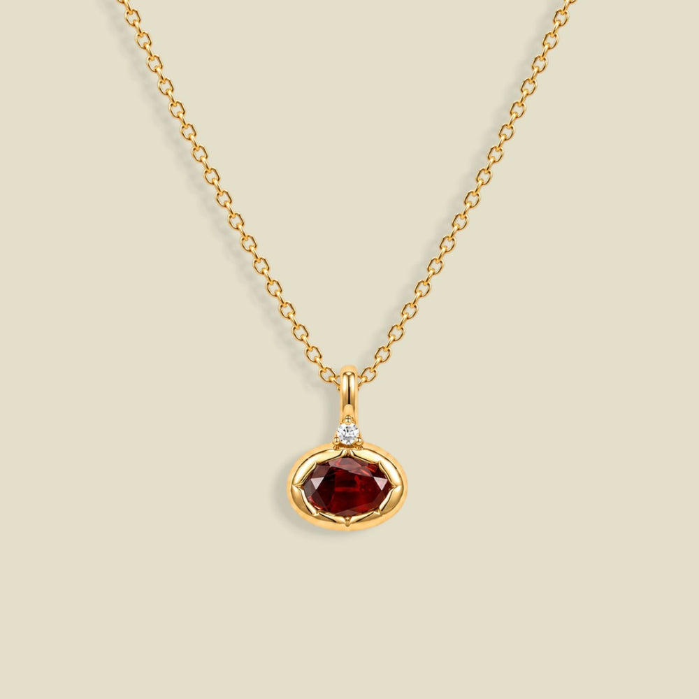 A gold chain necklace with a small oval pendant featuring a faceted dark red gemstone and a tiny clear accent stone above it, shown on a light beige background.