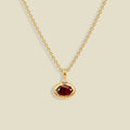A gold chain necklace with a small oval pendant featuring a faceted dark red gemstone and a tiny clear accent stone above it, shown on a light beige background.