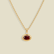 A gold chain necklace with a small oval pendant featuring a faceted dark red gemstone and a tiny clear accent stone above it, shown on a light beige background.