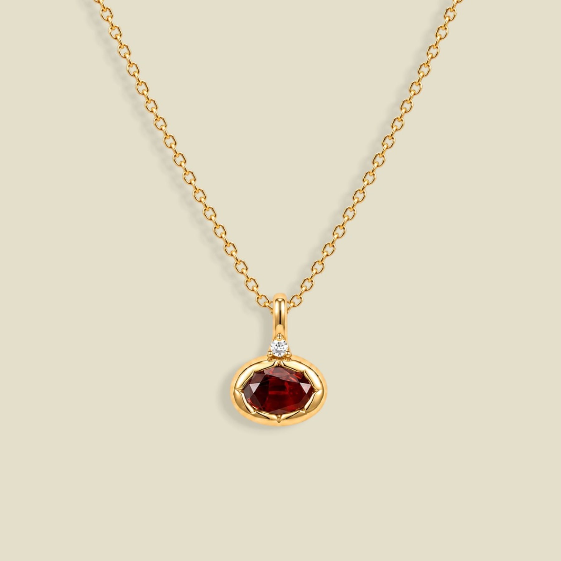 A gold chain necklace with a small oval pendant featuring a faceted dark red gemstone and a tiny clear accent stone above it, shown on a light beige background.
