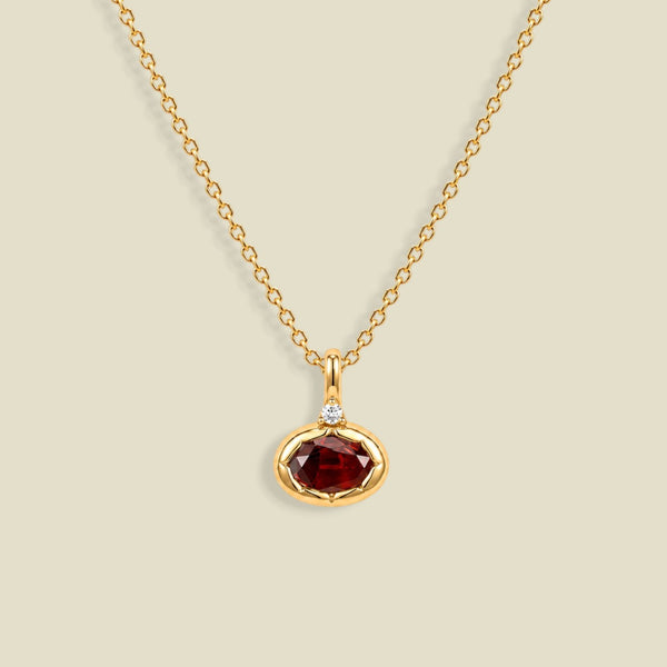 A gold chain necklace with a small oval pendant featuring a faceted dark red gemstone and a tiny clear accent stone above it, shown on a light beige background.