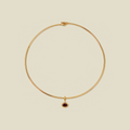 A delicate gold wire circle necklace with a small oval dark red gemstone pendant, displayed on a solid light beige background.