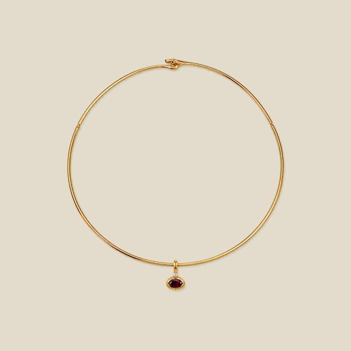 A delicate gold wire circle necklace with a small oval dark red gemstone pendant, displayed on a solid light beige background.